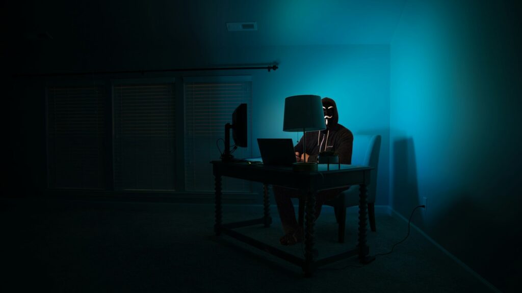 Persoon met masker achter laptop in donkere kamer symboliseert online anonimiteit en privacybescherming